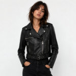 Woman wearing a premium black leather biker jacket featuring an asymmetrical zip closure, belted waist, lapel collar, and classic moto-inspired design.