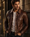 Handsome male model wearing a men's brown leather trucker jacket with snap-button closure, flap chest pockets, and classic vintage styling against an urban stone wall background.